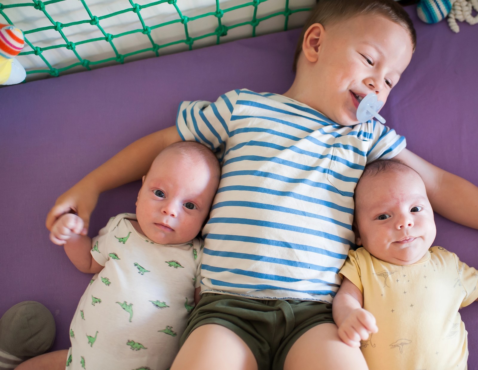 Älterer Bruder liegt im Kinderbett zwischen seinen beiden neugeborenen Zwillingsgeschwistern und hält ihre Hände